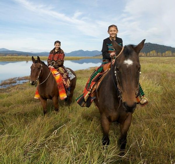 two tribal members riding horses near a river